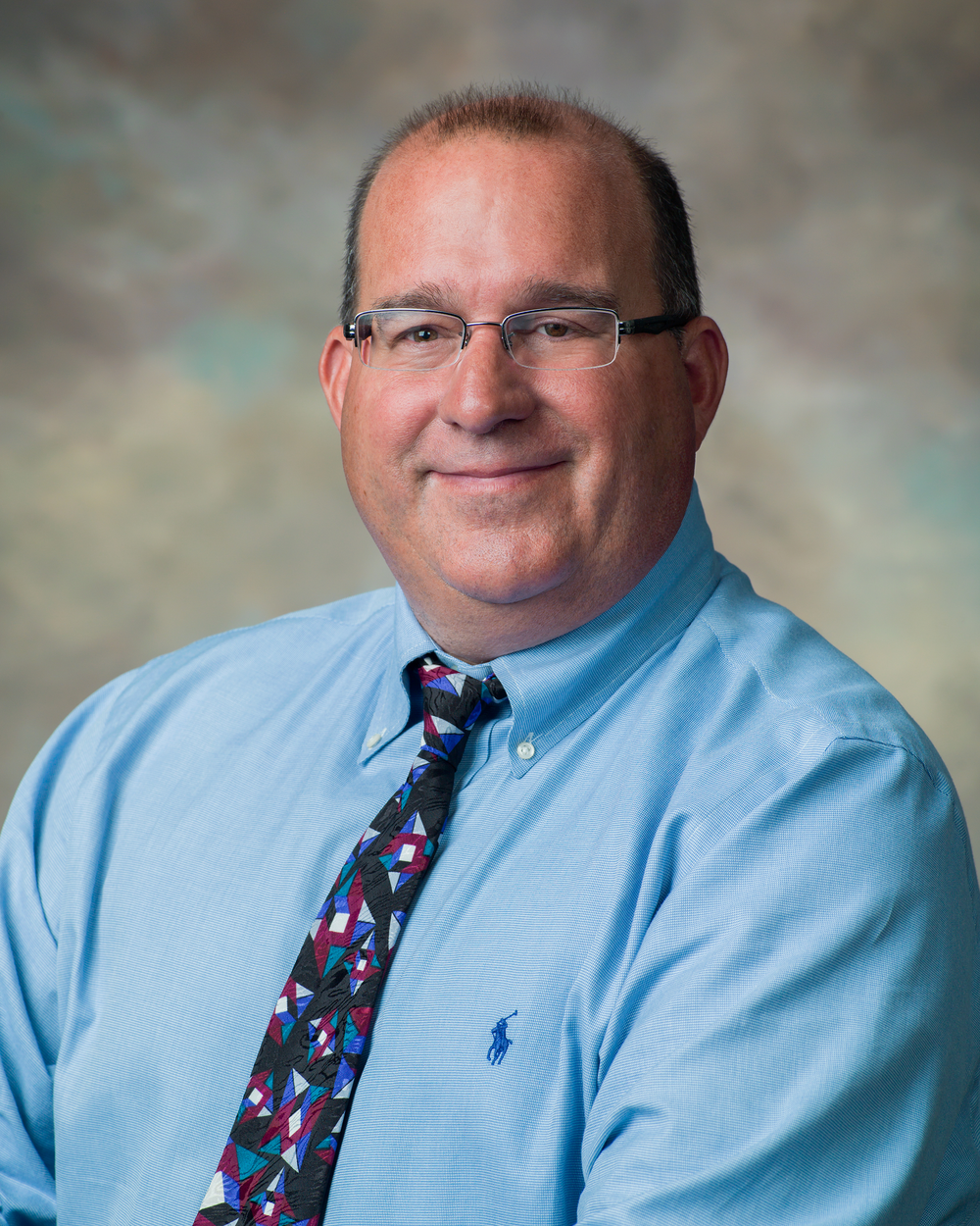 Official headshot of a man wearing glasses and a blue button down shirt.